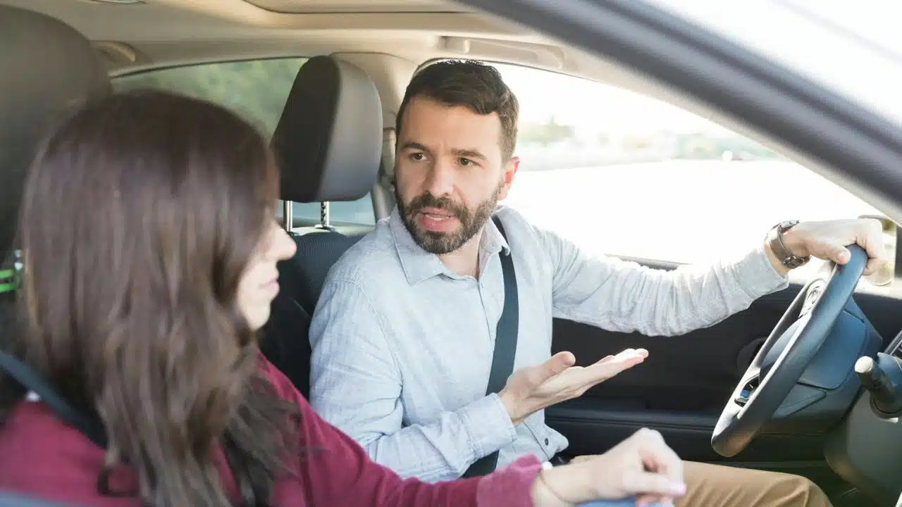 A man driving a car looks at a woman and gestures with an open palm.