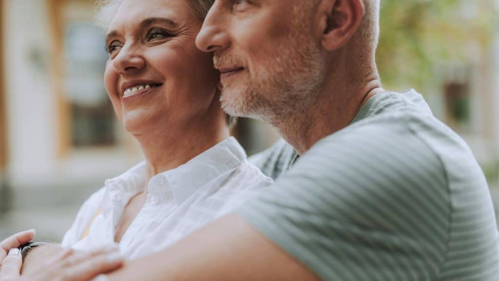 A smiling older couple standing close together and looking ahead.