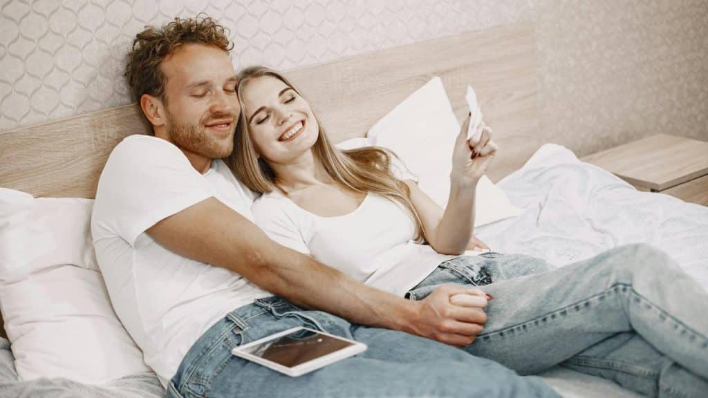 A happy couple sitting on a bed smiling and holding hands.
