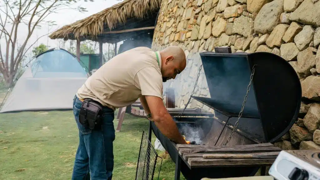 A man lighting a smoky grill outdoors near a tent and stone wall.