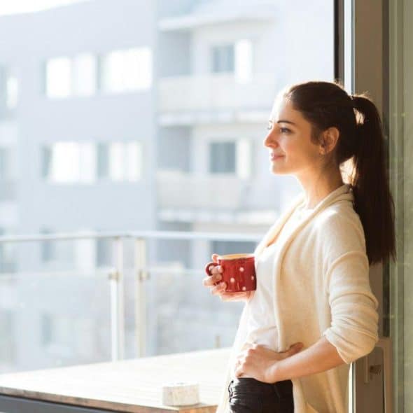 Woman holding a red polka-dot mug, looking out from a balcony on a sunny day.