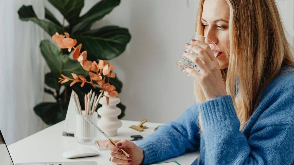 A woman in a blue sweater drinking water while writing at a desk.