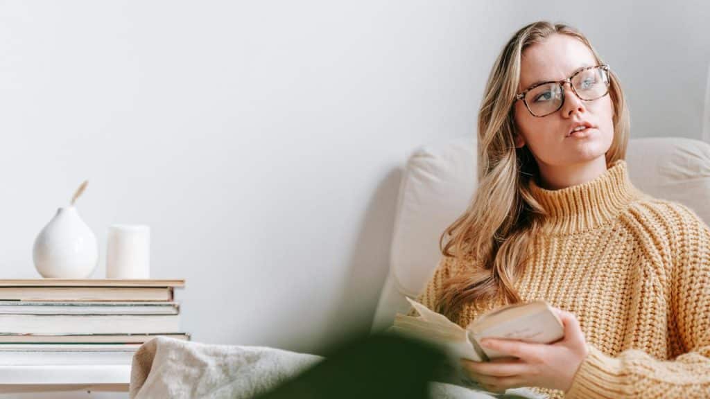 A woman wearing glasses and a sweater reading a book while sitting in a chair.