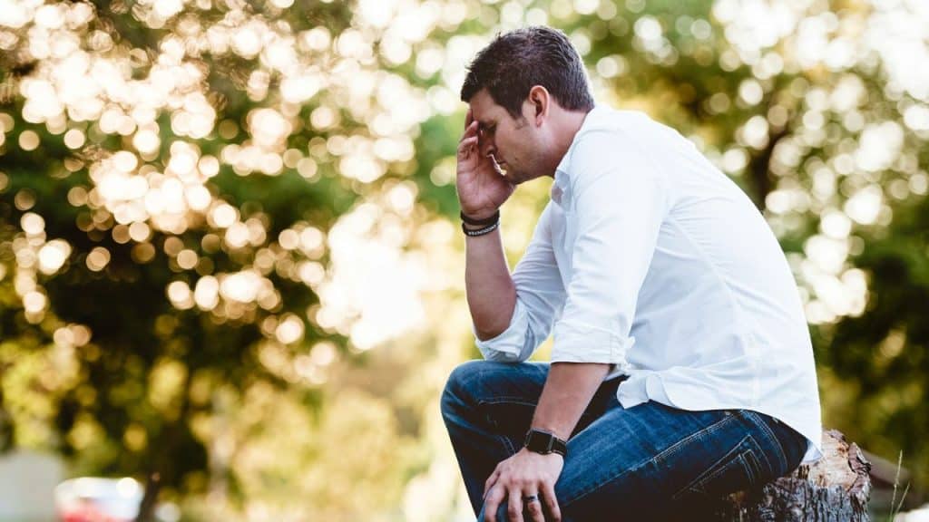 A man in a white shirt sits outside on a log with his hand on his forehead.