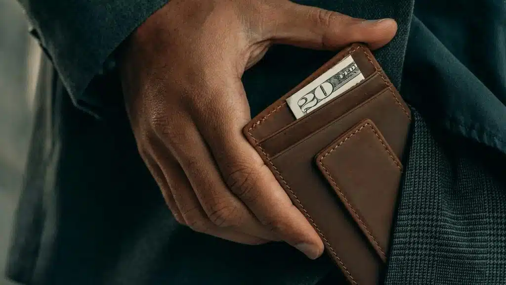 A person holding a brown wallet with a $20 bill visible.