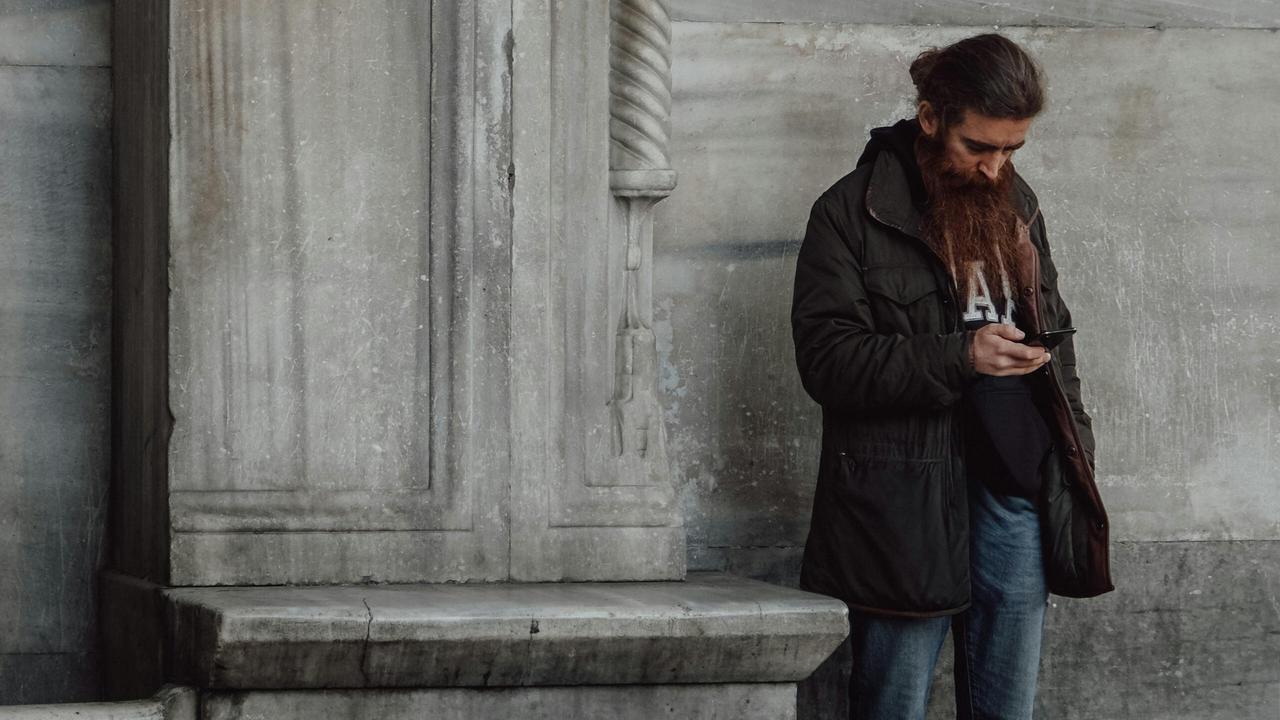 A bearded person standing outdoors, looking at a phone near a stone wall.