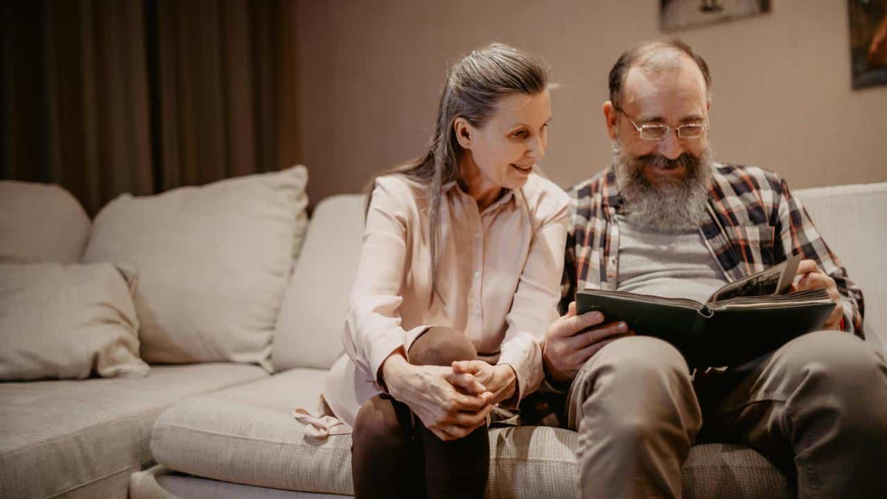 A senior couple sitting on a couch, looking at a photo album together.