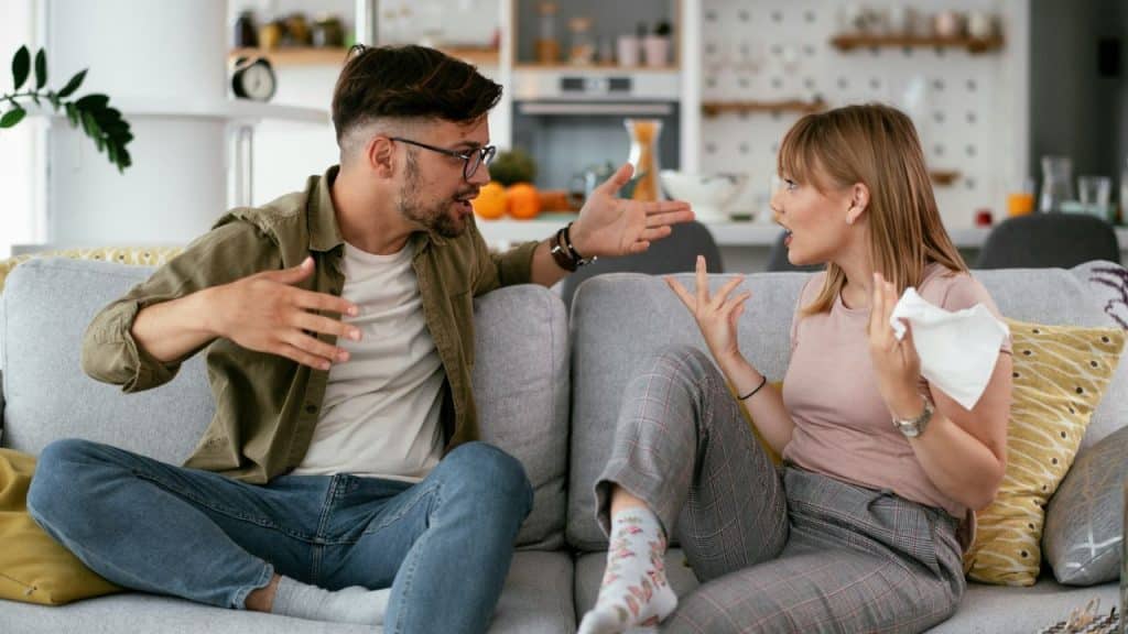 A man and woman sit on a grey sofa, gesturing with their hands while talking intensely.