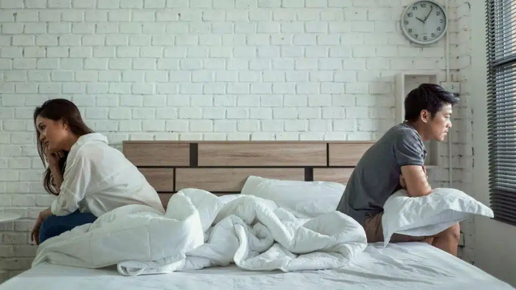 A man and woman sit on opposite sides of a bed, looking away from each other.