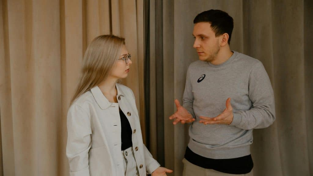 A man in a grey sweatshirt gestures with both hands toward a blonde woman.