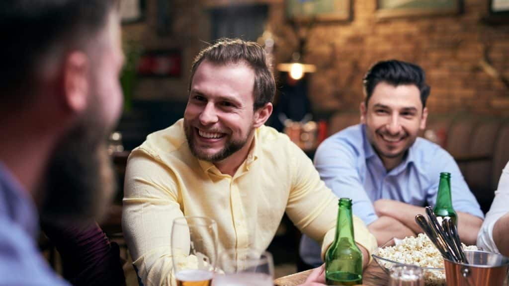Three men sit at a wooden bar table with drinks and a bowl of popcorn.