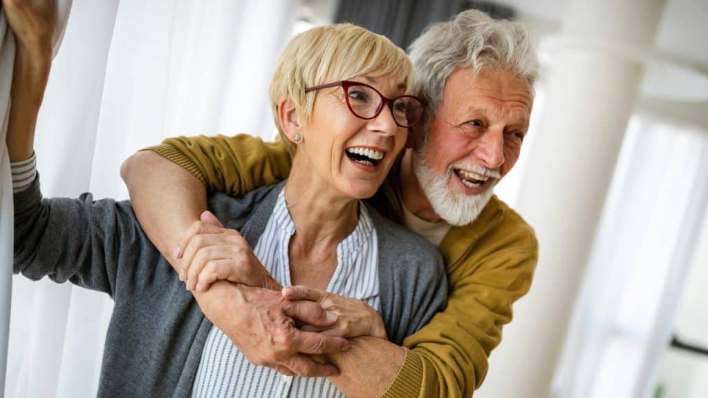 An older man with a grey beard hugs a smiling woman wearing red-framed glasses.