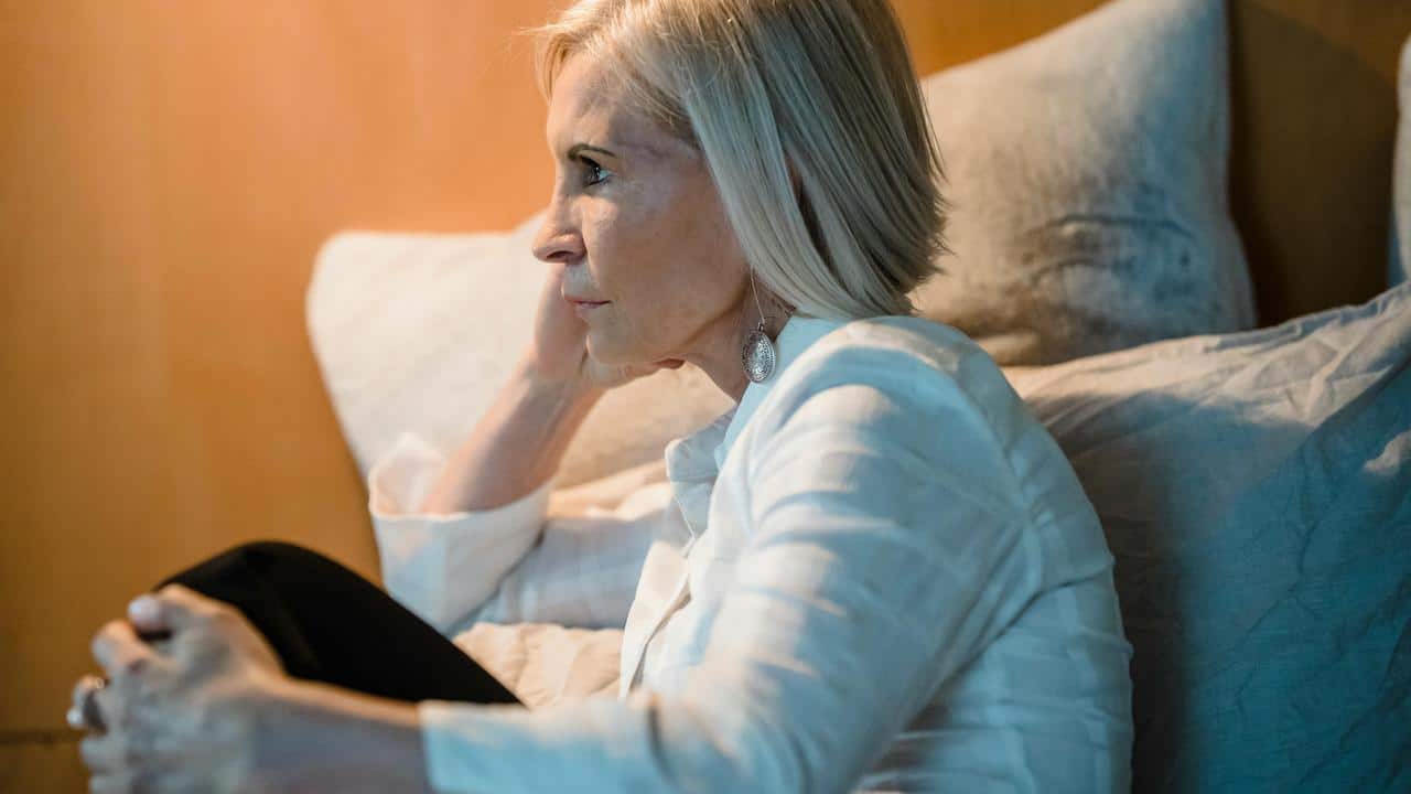A thoughtful older woman sitting on a couch with her hand resting against her head.