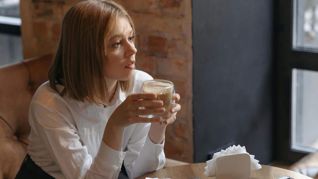 A woman sits by a window holding a glass cup of coffee in a cozy café.