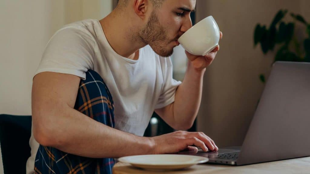 A man drinking from a cup while using a laptop at a table.