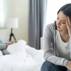 A distressed woman is sitting on the edge of a bed with her head in her hand, while a man sits turned away from her in the background.