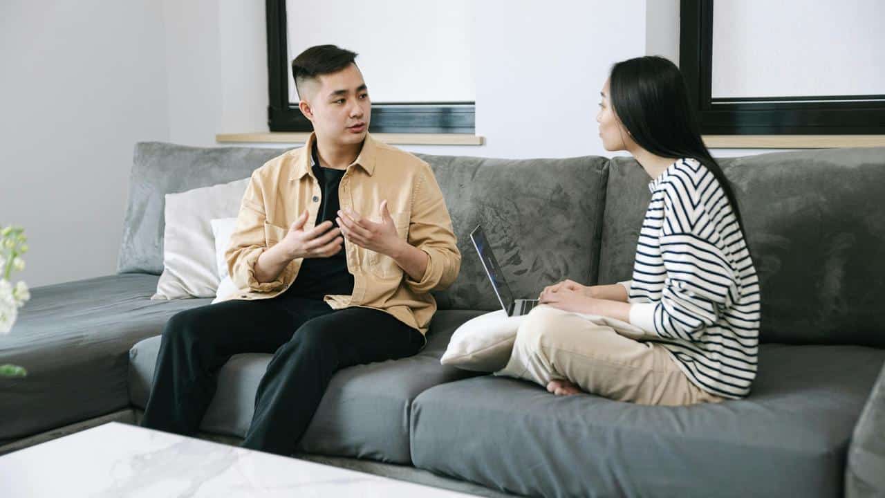 A man and a woman sitting on a couch, talking while using a laptop.