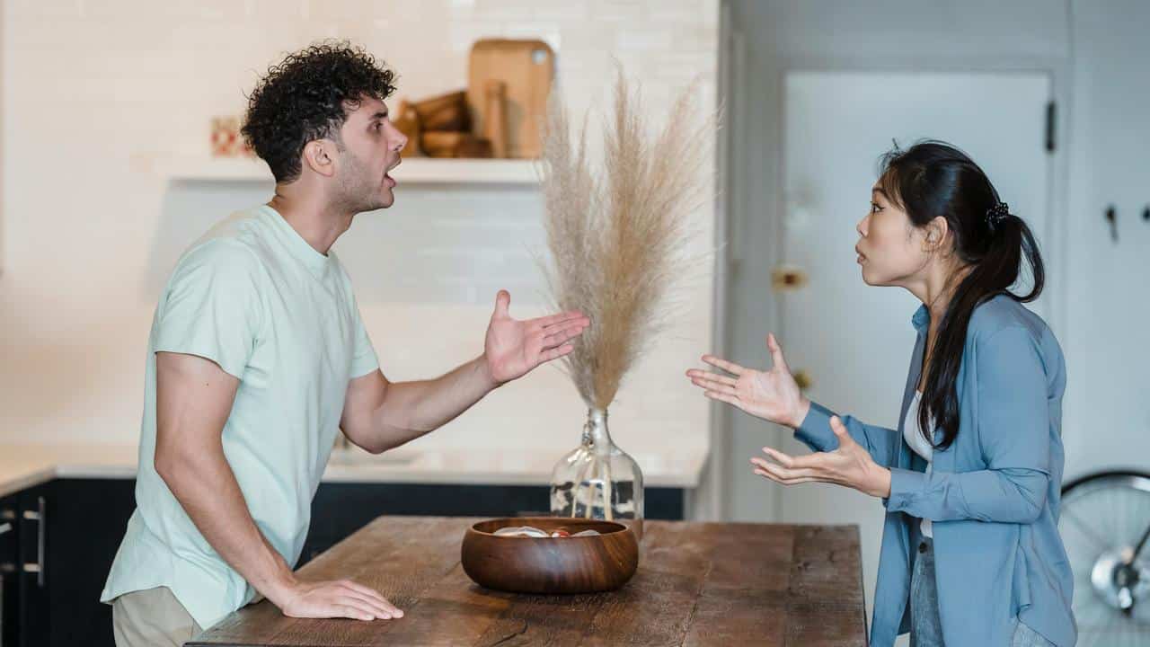 A man and a woman arguing across a table in a kitchen.