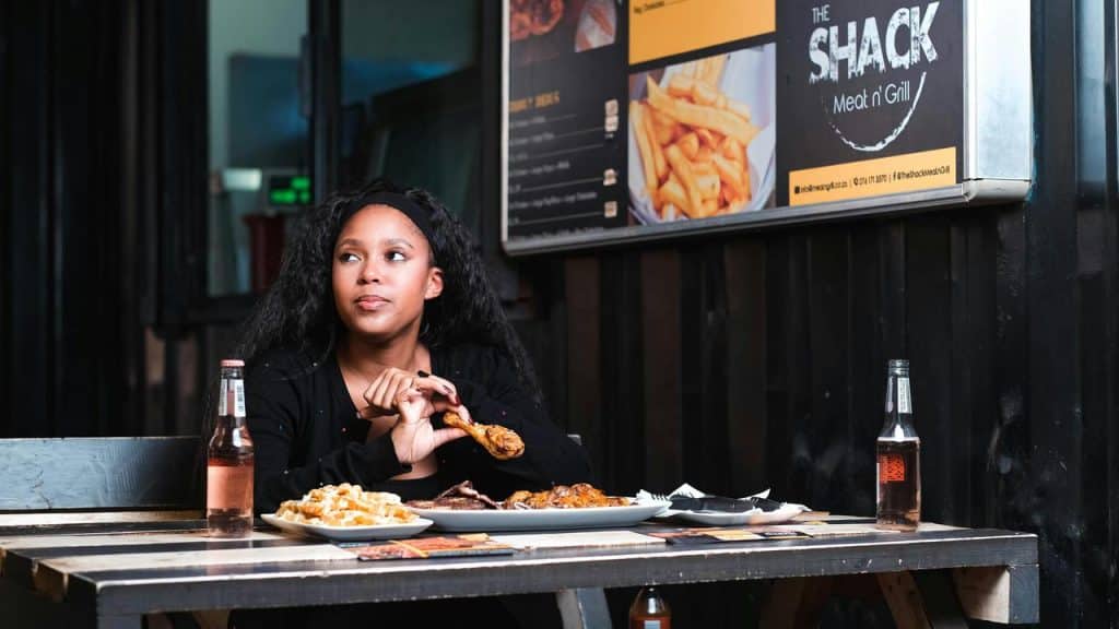 A woman sitting at an outdoor table eating a meal and holding a chicken drumstick.