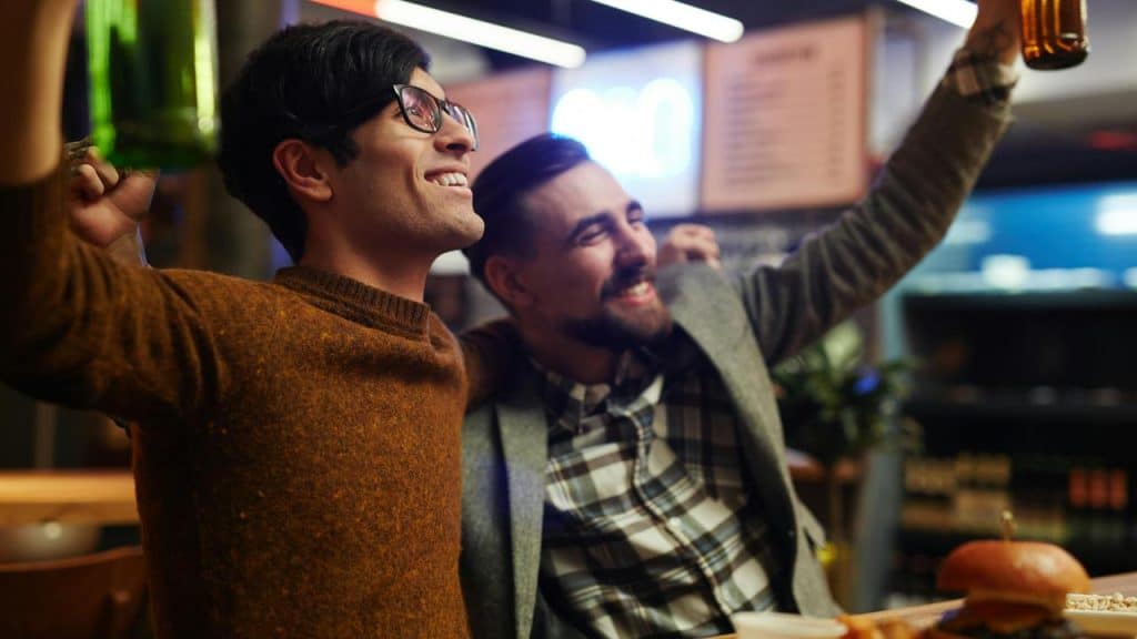A pair of men smiling and raising drinks while sitting together in a lively restaurant.