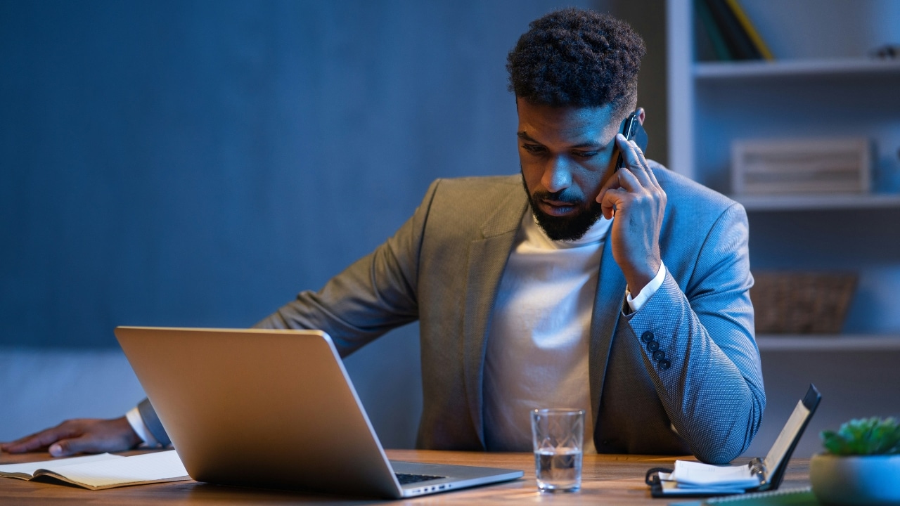 A man in a blazer sits at a desk with a laptop while talking on a cellphone.