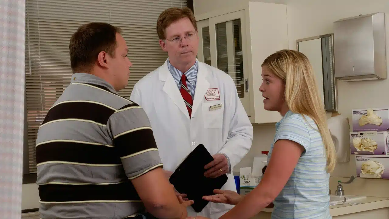 A doctor in a white coat talks to a man and woman in an office.