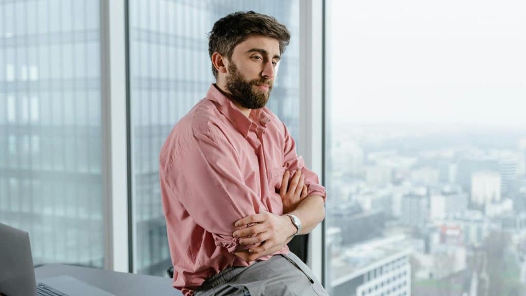 A man standing with arms crossed by a window, looking contemplative over a city view.