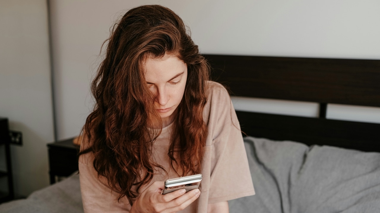 A woman with long wavy hair sits on a bed looking down at her smartphone.