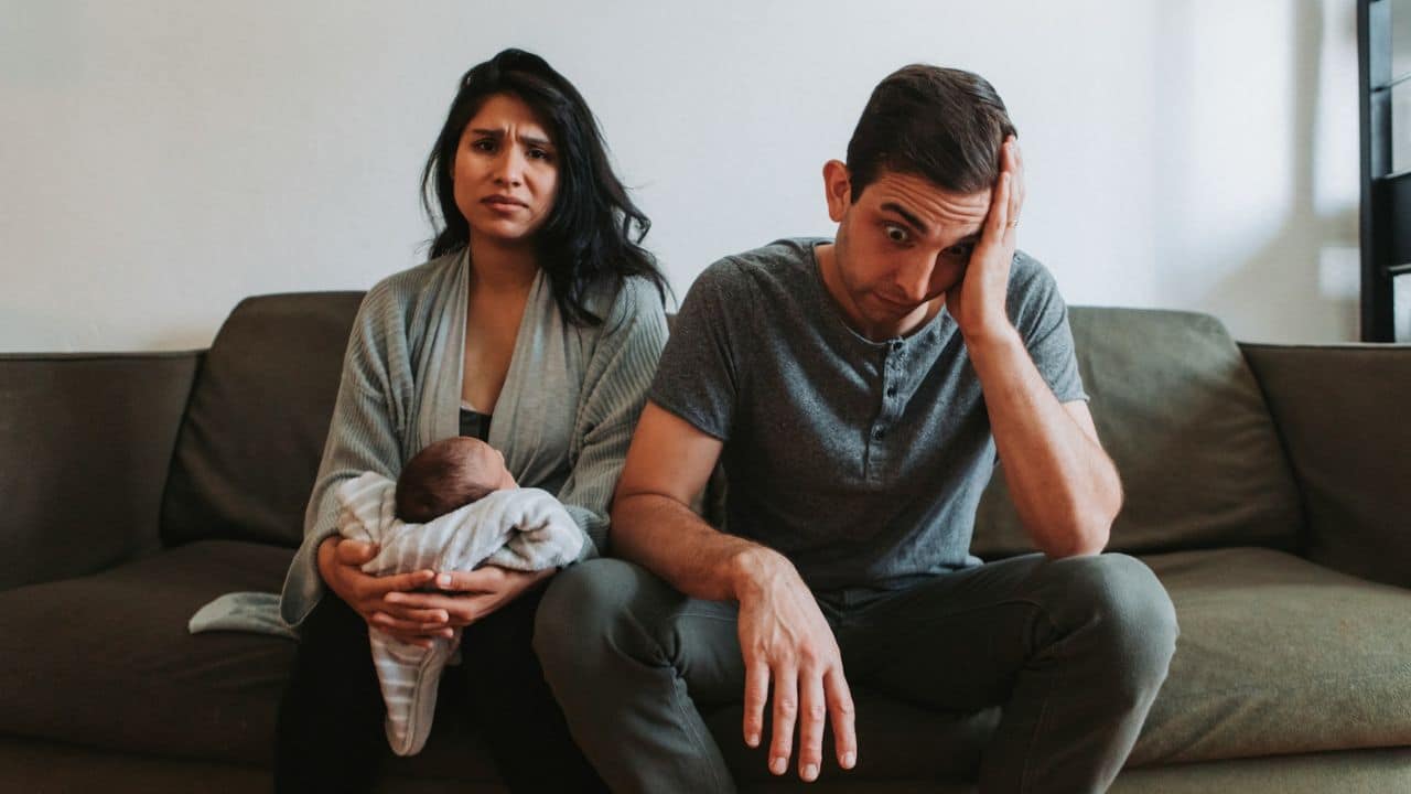 A woman holds a baby while a man beside her sits with his hand on his head.