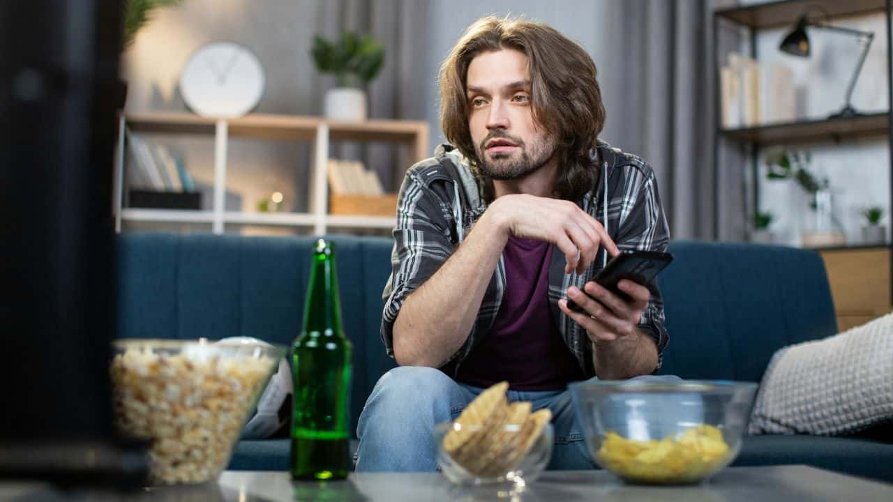 A man sits on a couch with a phone, surrounded by snacks and a green bottle.