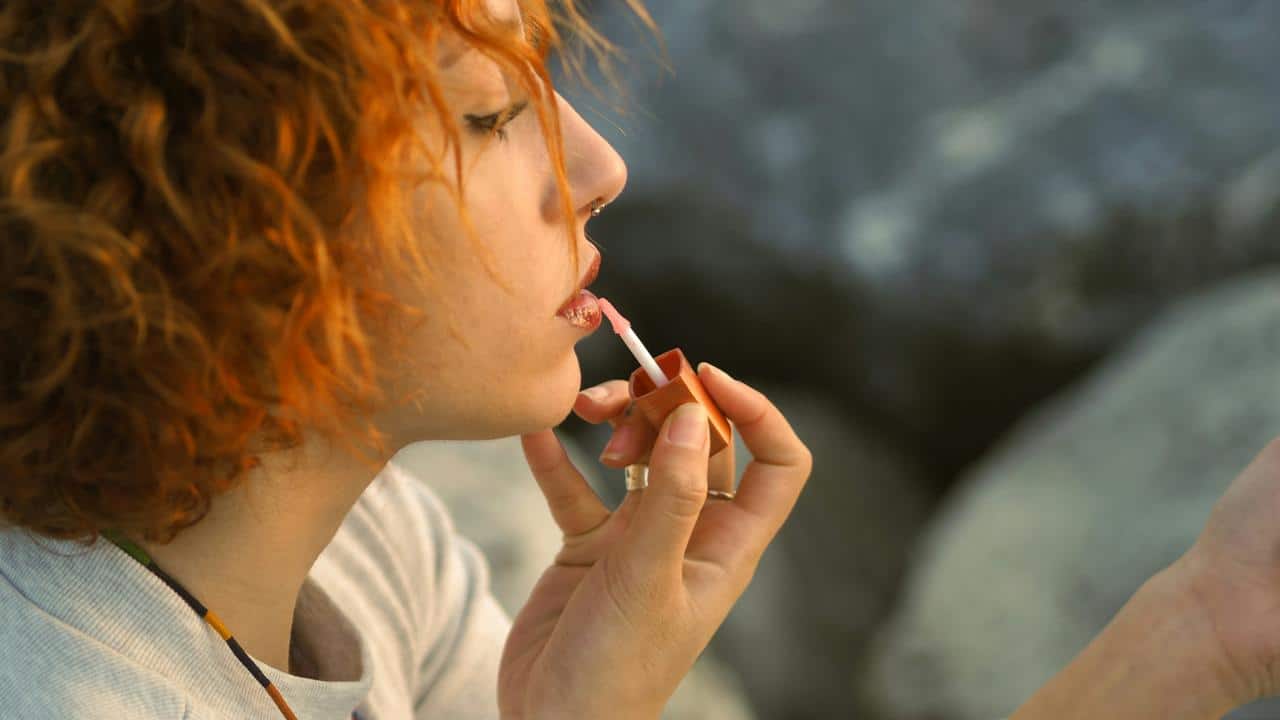 A woman applying lip gloss while looking at her reflection outdoors.