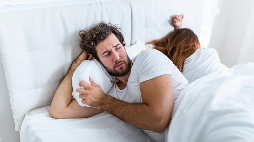 A man is lying in bed looking worried while a woman sleeps next to him with her back turned.
