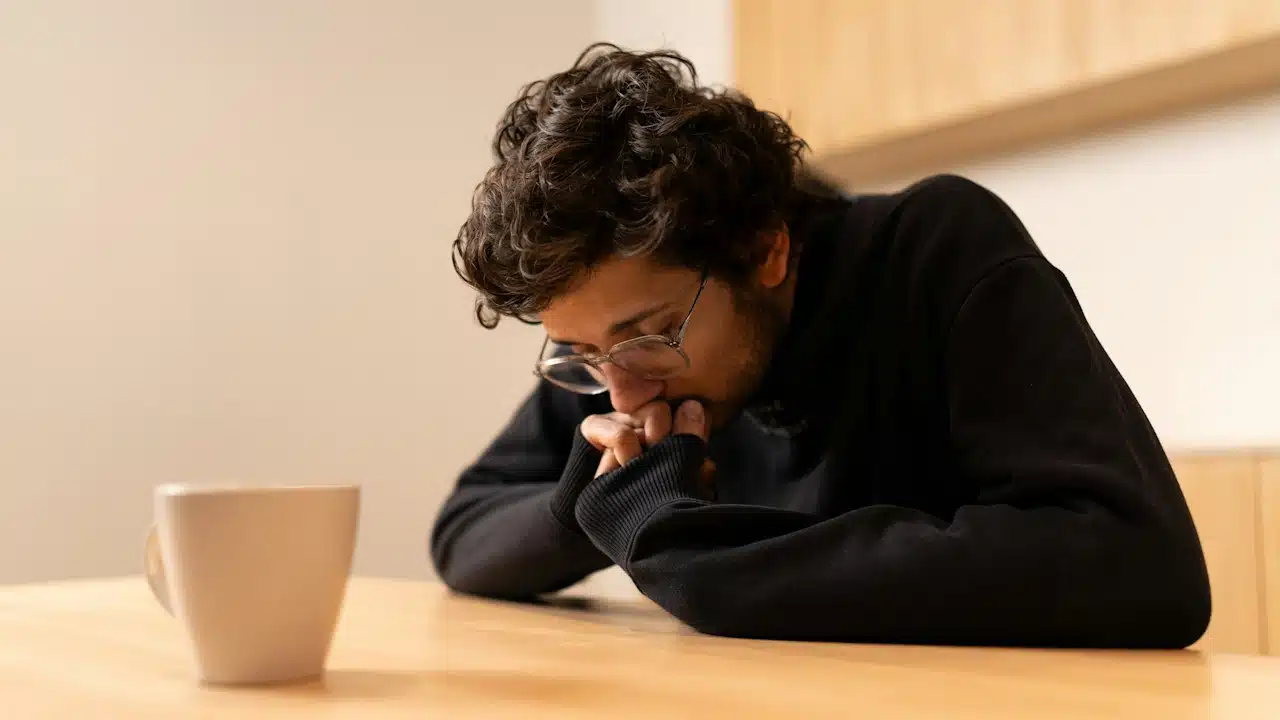 A man with glasses sitting at a table, looking down with a pensive expression, next to a white mug.