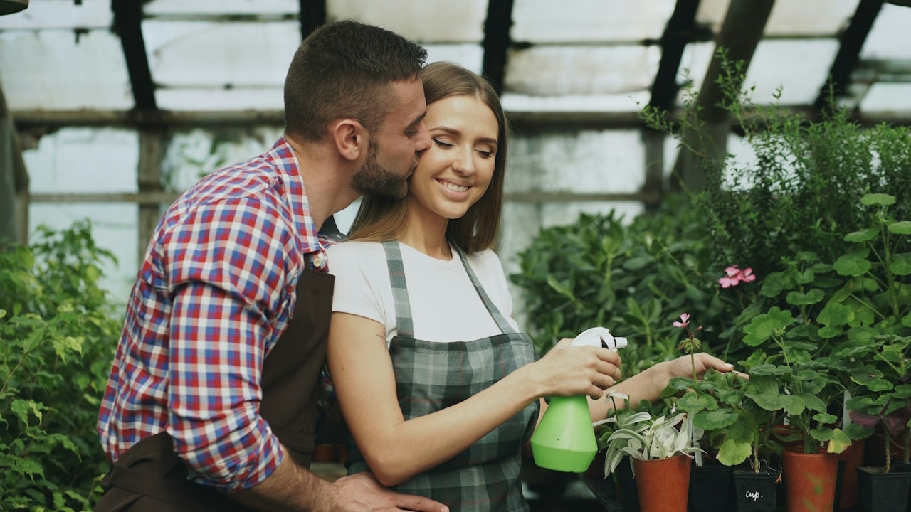 A man kissing a woman who’s tending to some plants in a greenhouse
