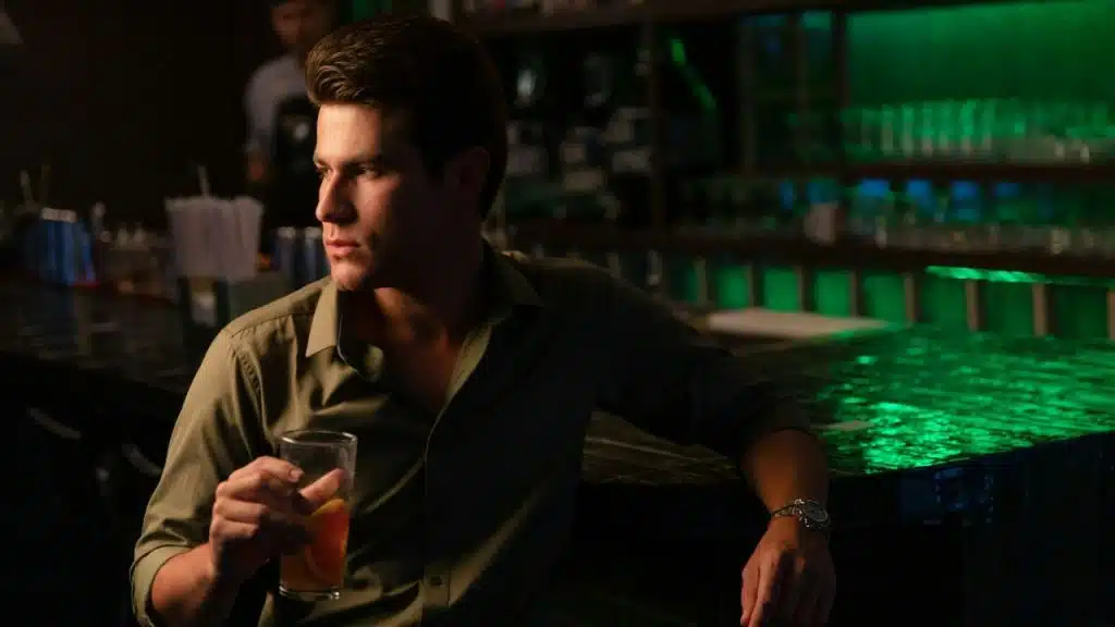 A man in a green shirt sits at a dimly lit bar holding a drink.
