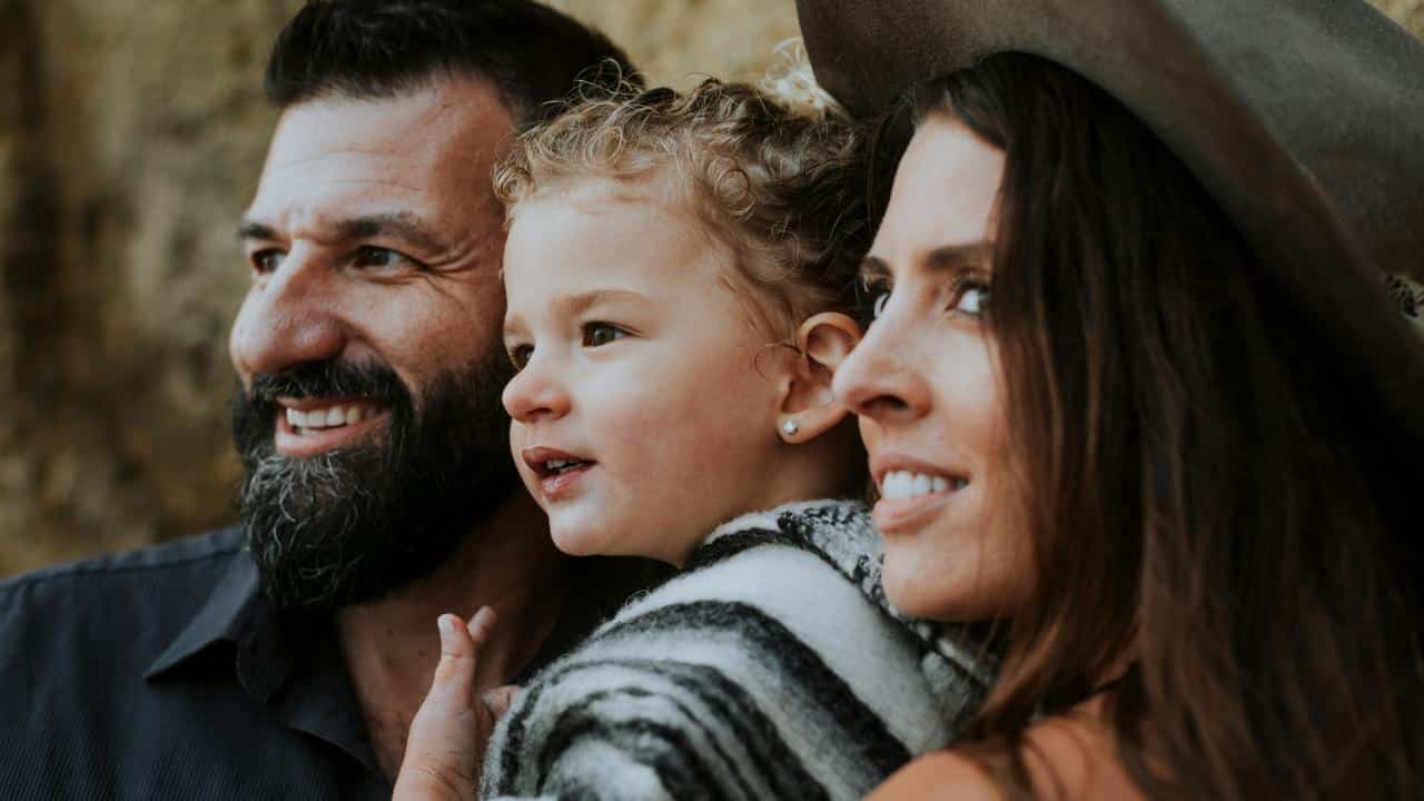 A close-up of two adults and a young child smiling together outdoors.