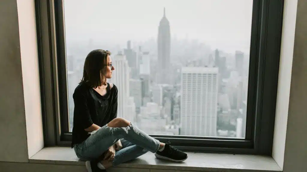 A woman sits by a large window, gazing out at a city skyline with tall buildings in the distance.