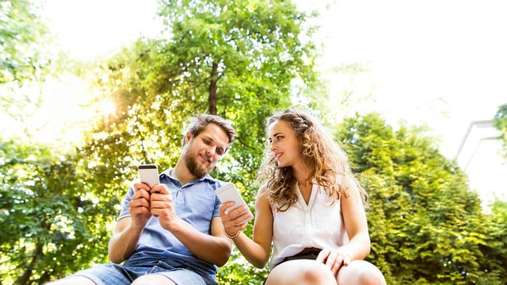 A couple sitting outdoors smiling while looking at their phones.