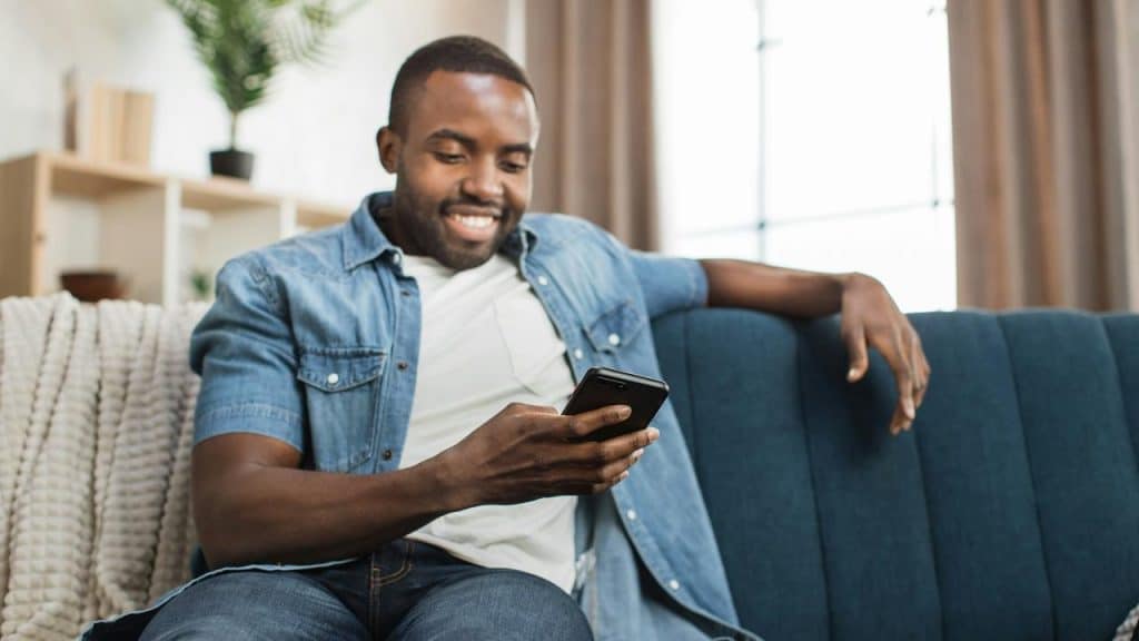 A man sitting on a couch smiling while looking at his smartphone.
