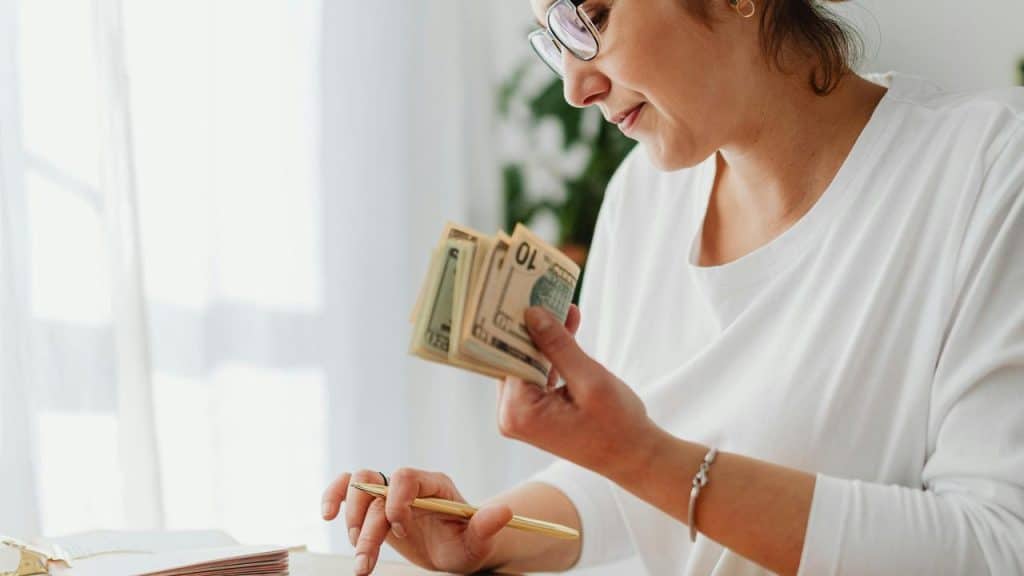 A woman counts paper money while writing at a table near a window.