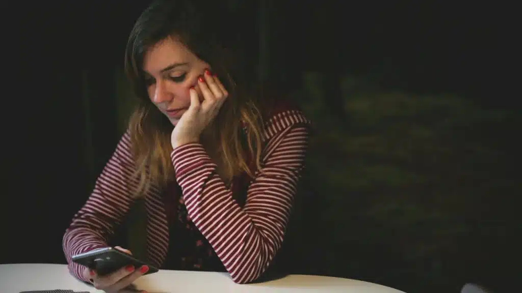 A young woman with her chin resting on her hand looking sadly at her smartphone