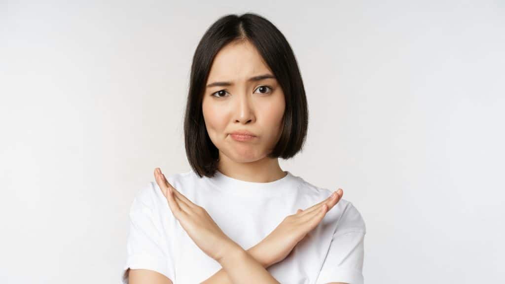 A woman with short dark hair in a white shirt makes an X with her arms.