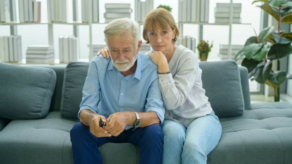 An older woman rests her chin on her hand while sitting beside a somber man.