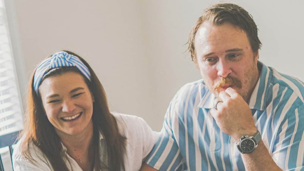 A man with a mustache and a woman wearing a blue striped headband laugh together.
