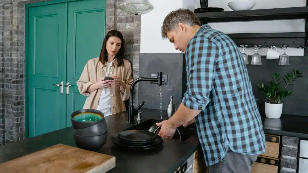 A man washes dishes in a kitchen sink while a woman leans nearby using a phone.