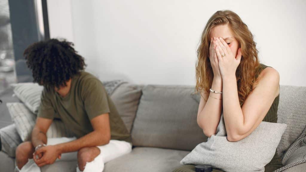 A woman sits on a sofa covering her face while a man sits nearby looking away.