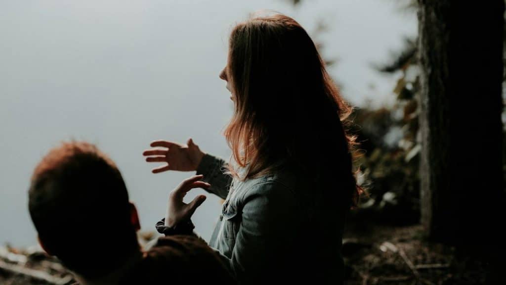 A woman gesturing with her hands while talking to a man outdoors by a foggy lake.