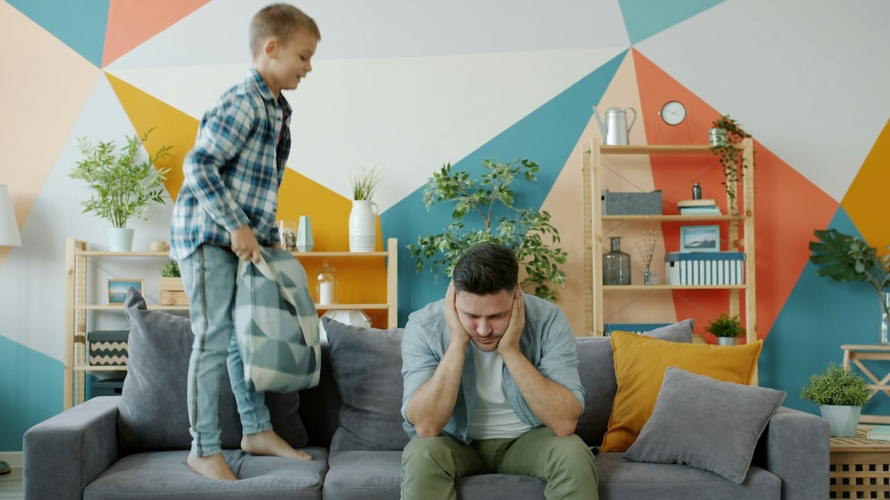 An annoyed man sitting on the sofa with his head in his hands while a kid stands beside him with a pillow.