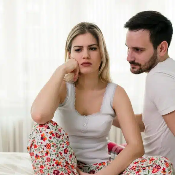 A sad woman is sitting on a bed, and her husband is looking at her while sitting next to her.