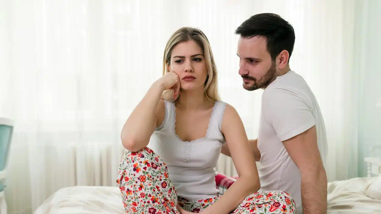 A sad woman is sitting on a bed, and her husband is looking at her while sitting next to her.
